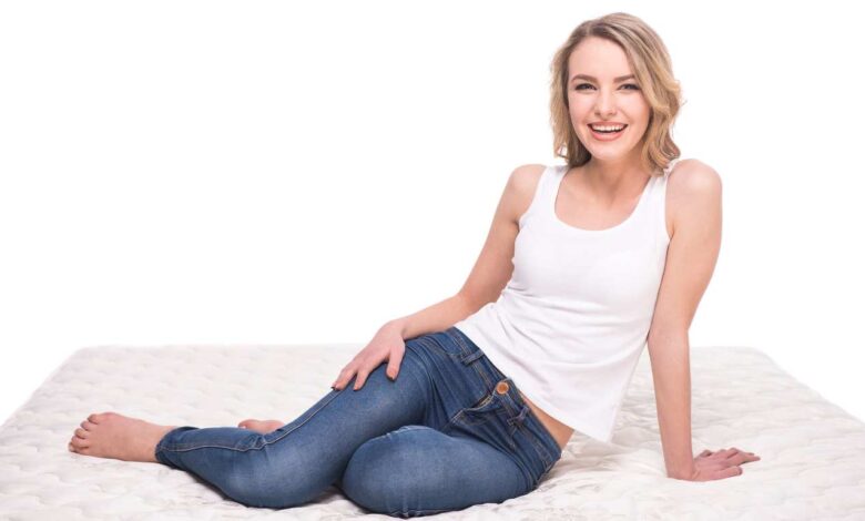 a girl in white shirt and blue jeans pant sitting on a white cheap mattress smiling at a camera