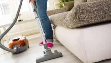a person in lite blue jeans pant and pink shoe standing beside gray and white sofa using canister vacuum on cream coloured floor.
