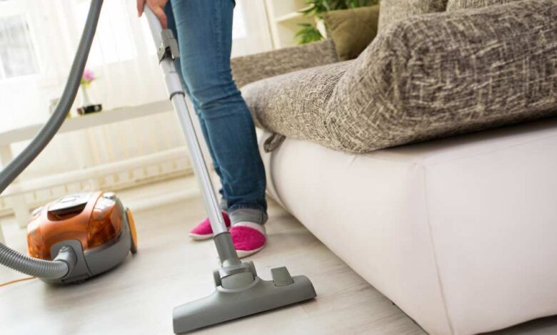 a person in lite blue jeans pant and pink shoe standing beside gray and white sofa using canister vacuum on cream coloured floor.